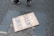 A cardboard sign with the message 'TO BE SILENT IS TO BE COMPLICIT' written in bold blue and white letters lies on the asphalt. There are a pair of feet in sneakers visible above the sign, suggesting someone passing by or participating in a protest.