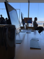 A sleek modern workspace features several large computer monitors and keyboards arranged neatly on a wooden table. Natural light pours in through wide glass windows, creating a spacious and inviting atmosphere. In the background, there are a few people, with one person seated and looking out through the windows, contributing to a sense of calm and focus.