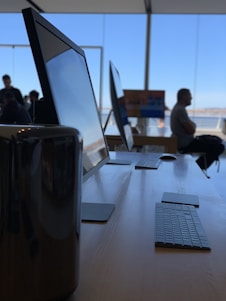 A sleek modern workspace features several large computer monitors and keyboards arranged neatly on a wooden table. Natural light pours in through wide glass windows, creating a spacious and inviting atmosphere. In the background, there are a few people, with one person seated and looking out through the windows, contributing to a sense of calm and focus.
