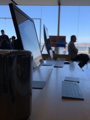 A sleek modern workspace features several large computer monitors and keyboards arranged neatly on a wooden table. Natural light pours in through wide glass windows, creating a spacious and inviting atmosphere. In the background, there are a few people, with one person seated and looking out through the windows, contributing to a sense of calm and focus.