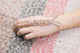 A hand adorned with intricate henna design featuring floral patterns and dotted lines. The background consists of a textured, multi-colored fabric.