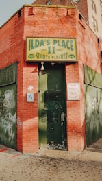 A brick building with an aged appearance and some graffiti. The entrance features a dark green, slightly worn door beneath a sign reading 'ILDA'S PLACE II, SPORTS CABARET'. A letter 'A' health grade sign and a notice are also visible next to the door.