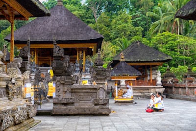 An intricately designed temple with ornate stone carvings and traditional thatched roofs surrounded by lush greenery. A group of people in traditional attire are sitting on the temple grounds, engaging in a ceremonial activity.