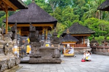 An intricately designed temple with ornate stone carvings and traditional thatched roofs surrounded by lush greenery. A group of people in traditional attire are sitting on the temple grounds, engaging in a ceremonial activity.