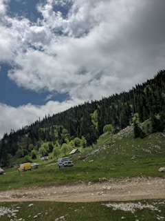 A lush mountainside covered with dense, evergreen forest under a partly cloudy sky. On the grassy slope, there are several vehicles, including a yellow van and a dark-colored SUV, next to a small, rustic wooden cabin. The scene is serene and isolated, evoking a sense of tranquility in nature.