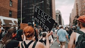 A crowded street scene with people participating in a protest. One person holds up a sign reading 'Black Lives Matter' in bold letters, while another sign reads 'White Silence is Violence'. The urban setting features tall buildings on either side of the street, and the participants appear engaged and focused.