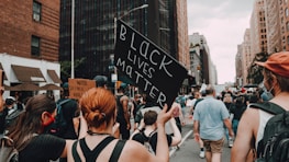 A crowded street scene with people participating in a protest. One person holds up a sign reading 'Black Lives Matter' in bold letters, while another sign reads 'White Silence is Violence'. The urban setting features tall buildings on either side of the street, and the participants appear engaged and focused.