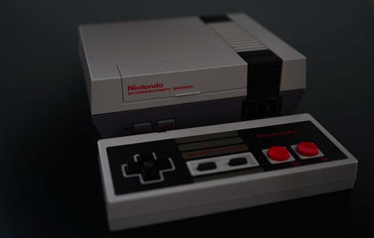 A classic gray gaming console with a matching controller on a dark surface. The console features a front-loading cartridge slot and two controller ports, while the controller has a D-pad and red buttons.