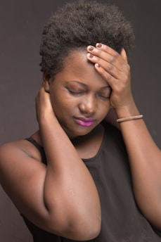 A person with closed eyes rests their hand on their forehead and another on their neck, conveying a sense of discomfort or stress. They have short curly hair, wear a sleeveless black top, and have brightly colored lipstick.