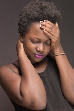A person with closed eyes rests their hand on their forehead and another on their neck, conveying a sense of discomfort or stress. They have short curly hair, wear a sleeveless black top, and have brightly colored lipstick.