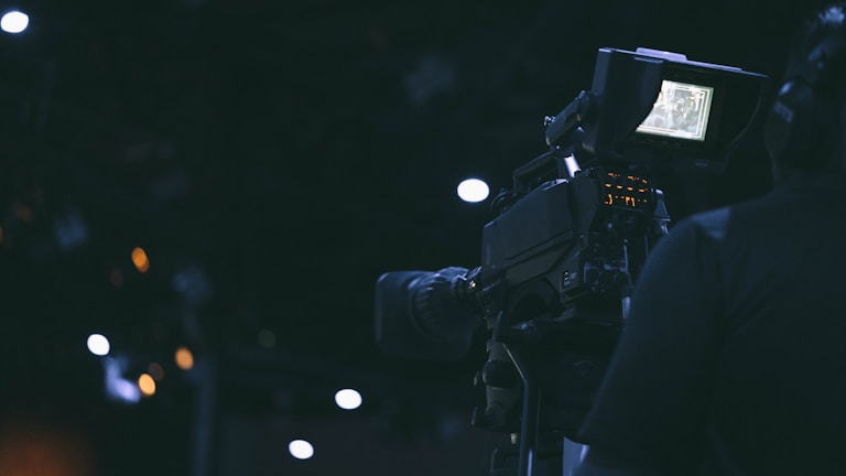 A film camera is being operated by a person in a dimly lit environment. The camera's screen is illuminated, displaying some details, while the surroundings are characterized by dark shades and a few out-of-focus lights.