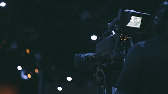 A film camera is being operated by a person in a dimly lit environment. The camera's screen is illuminated, displaying some details, while the surroundings are characterized by dark shades and a few out-of-focus lights.