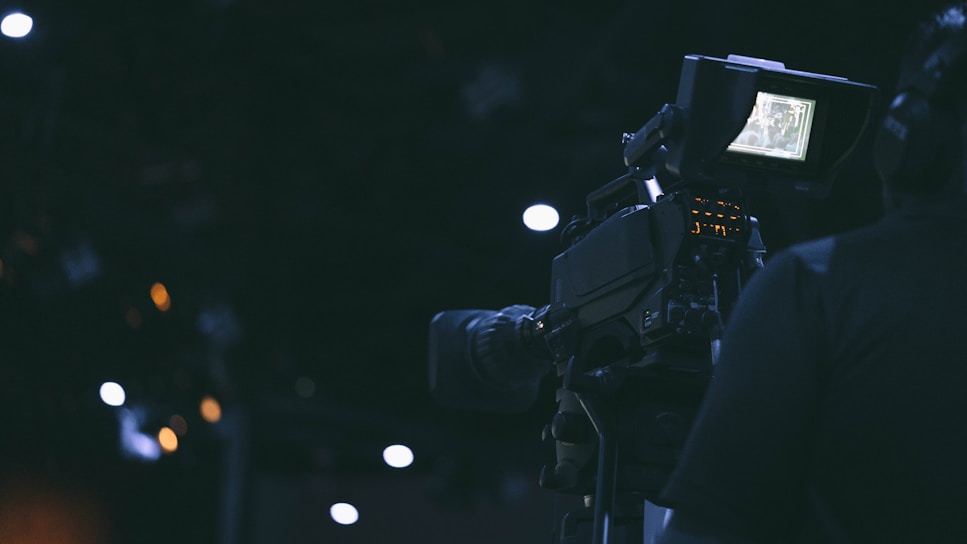A film camera is being operated by a person in a dimly lit environment. The camera's screen is illuminated, displaying some details, while the surroundings are characterized by dark shades and a few out-of-focus lights.