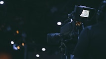 A film camera is being operated by a person in a dimly lit environment. The camera's screen is illuminated, displaying some details, while the surroundings are characterized by dark shades and a few out-of-focus lights.