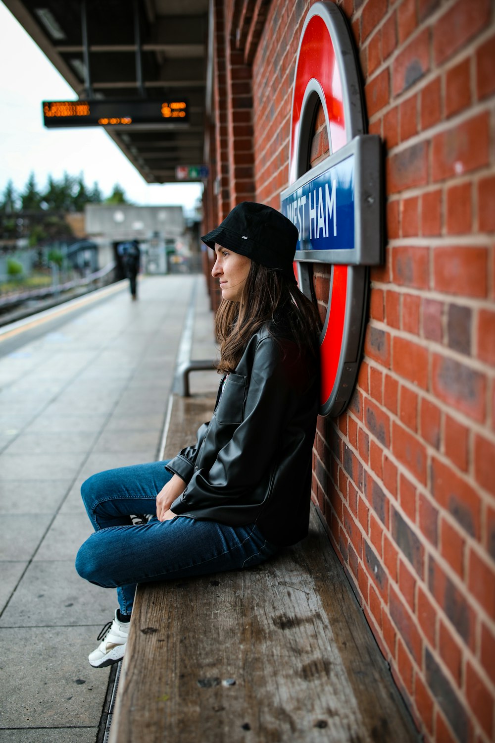 Donna in giacca nera e jeans blu in denim che si siede sul muro di mattoni rosso durante il giorno