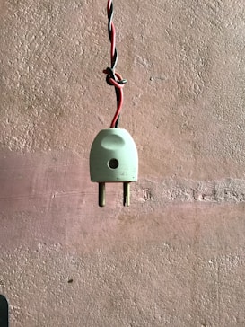 A white two-pin electrical plug with red and black twisted wires hangs against a textured wall. The wall features a horizontal band of lighter color across the middle.