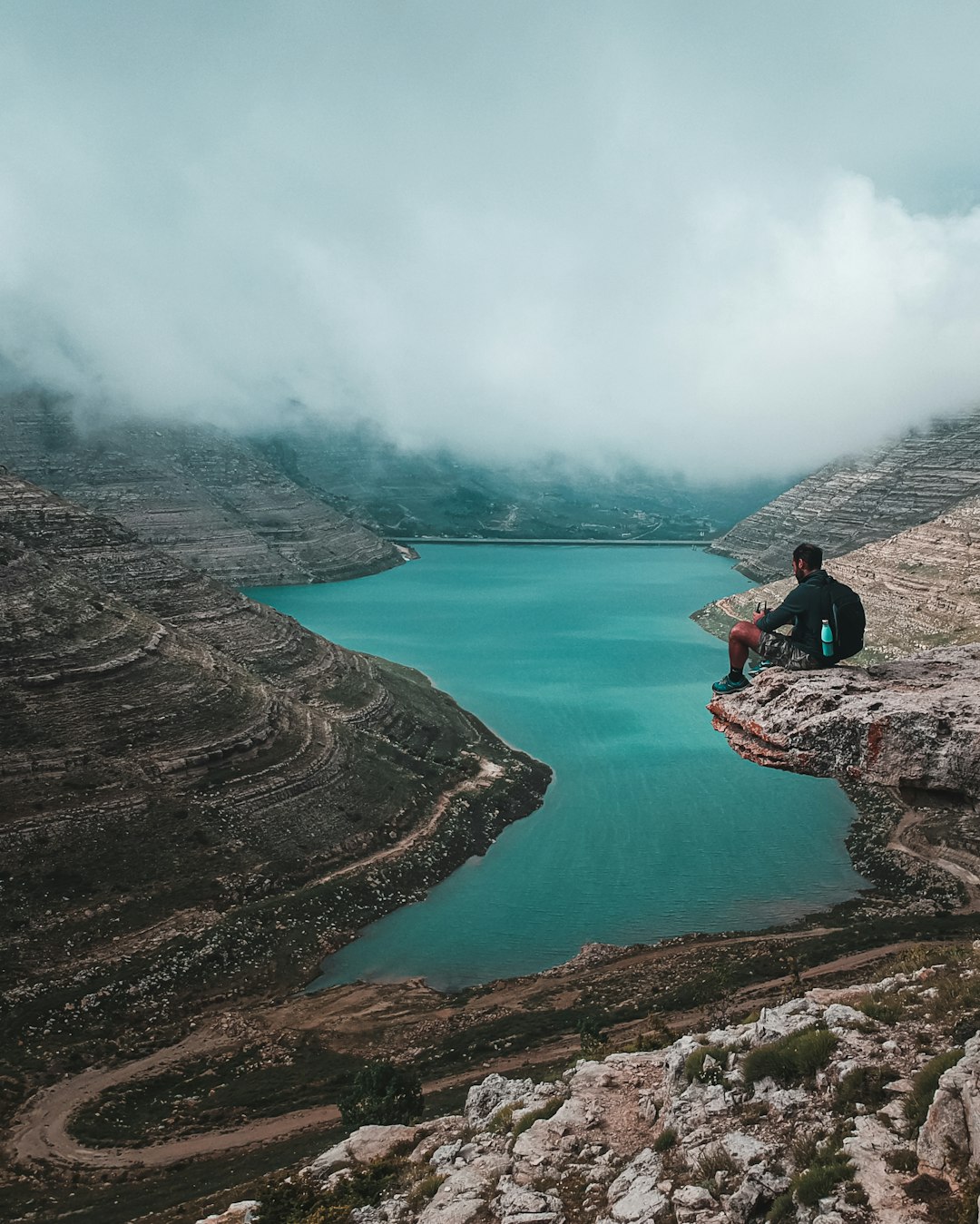 man in black jacket sitting on rock near lake during daytime