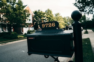 A close-up of a black mailbox with the number 9726 displayed on top. The mailbox is positioned along a suburban street with trees and houses in the background. The lighting suggests it is either early evening or late afternoon.