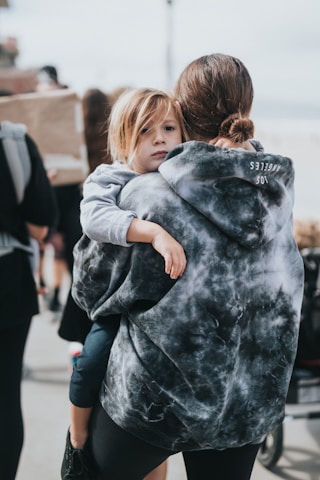 A child with blonde hair is being carried by a person wearing a dark, tie-dye hoodie. The child looks towards the camera with a neutral expression. There are other people walking in the background holding signs, suggesting they are part of a group or event.