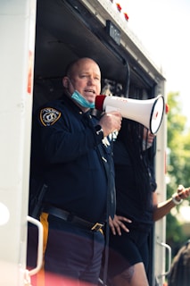 A uniformed police officer is standing in the doorway of a vehicle, holding a megaphone. The officer is wearing a mask under the chin and appears to be addressing a crowd. Another person partially in view stands nearby, resting against the vehicle.