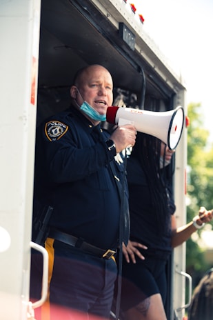 A uniformed police officer is standing in the doorway of a vehicle, holding a megaphone. The officer is wearing a mask under the chin and appears to be addressing a crowd. Another person partially in view stands nearby, resting against the vehicle.