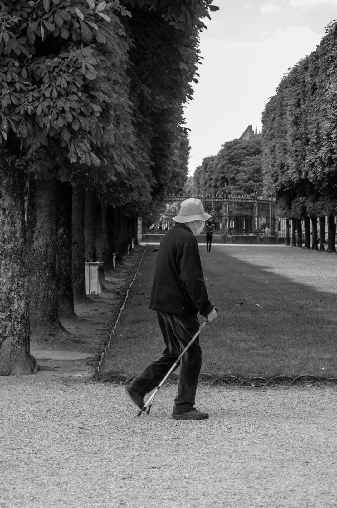 grayscale photo of man in black coat and hat walking on pathway