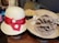 Two stylish straw hats displayed on a wooden surface, featuring distinct decorative bands. The hat on the left has a prominent red ribbon with two white buttons, while the hat on the right has a large brown fabric flower and lace detailing.