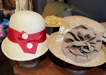 Two stylish straw hats displayed on a wooden surface, featuring distinct decorative bands. The hat on the left has a prominent red ribbon with two white buttons, while the hat on the right has a large brown fabric flower and lace detailing.