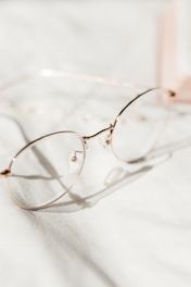 A pair of round, gold-rimmed eyeglasses is positioned on a soft, white fabric surface. The light creates gentle shadows and highlights the delicate structure of the glasses.