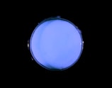 A round petri dish containing a light blue substance, set against a black background. The edges of the dish are barely visible, creating a glowing illusion.
