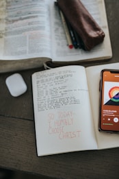 An open book with handwritten notes sits on a wooden table. Beside it is a smartphone displaying a music player app, and in front of the book is a notebook with handwritten words in red ink stating 'SO TODAY I HUMBLY CHOOSE CHRIST'. A leather pouch containing pens and an AirPods case are also present.