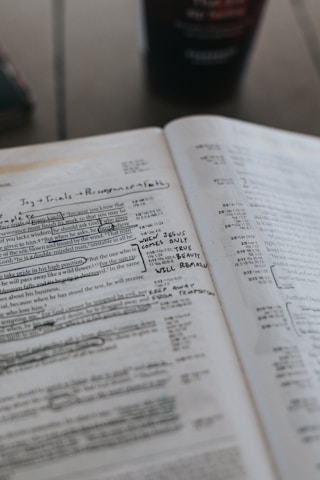 An open Bible with handwritten notes and underlined text. The pages are filled with annotations in pen, emphasizing certain passages. A blurred background suggests a cup is present on the table.