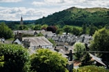 A quaint village nestled in a lush, green landscape is surrounded by trees and rolling hills in the background. The village features traditional architecture with stone and slate roofs. A tall church spire rises prominently among the buildings, adding to the idyllic, peaceful atmosphere.