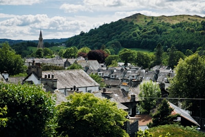 A quaint village nestled in a lush, green landscape is surrounded by trees and rolling hills in the background. The village features traditional architecture with stone and slate roofs. A tall church spire rises prominently among the buildings, adding to the idyllic, peaceful atmosphere.