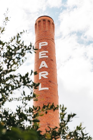 A tall, red brick chimney with the word 'PEARL' written vertically in white letters. The sky is partially cloudy, and green foliage frames the lower part of the image.