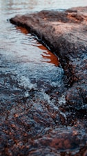 Water gently flows over a dark, textured rock surface with small bubbles. The scene captures the dynamic movement of the clear water and the contrasting texture of the rock.