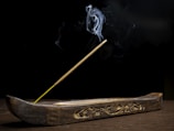 A wooden incense holder with intricate carvings holds a single incense stick. Smoke elegantly rises from the tip, creating delicate patterns against a dark background. The overall setting suggests simplicity and calmness.