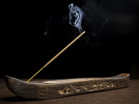 A wooden incense holder with intricate carvings holds a single incense stick. Smoke elegantly rises from the tip, creating delicate patterns against a dark background. The overall setting suggests simplicity and calmness.