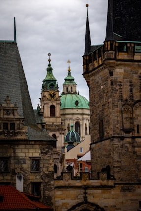 A view of historical buildings with ornate architecture. The central part of the image features a structure with green domes and gold accents, including a clock. Surrounding this are various stone buildings with intricate details and steep roofs, suggesting a European architectural style.