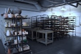 A room filled with metal shelving units holding numerous pottery and ceramic items in various stages of completion. The industrial ceiling with pipes and exposed ducts suggests a workshop or studio environment. The lighting is dim, casting shadows on the floor and racks.