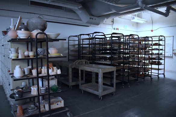 A room filled with metal shelving units holding numerous pottery and ceramic items in various stages of completion. The industrial ceiling with pipes and exposed ducts suggests a workshop or studio environment. The lighting is dim, casting shadows on the floor and racks.