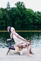 A person is seated on a purple portable massage chair at the edge of a body of water, covered with a patterned blanket, eyes closed and face slightly elevated. The background features lush green trees, and there is a small cloth and a bottle placed nearby on the ground.