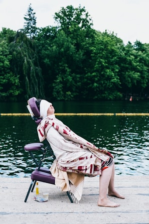 A person is seated on a purple portable massage chair at the edge of a body of water, covered with a patterned blanket, eyes closed and face slightly elevated. The background features lush green trees, and there is a small cloth and a bottle placed nearby on the ground.