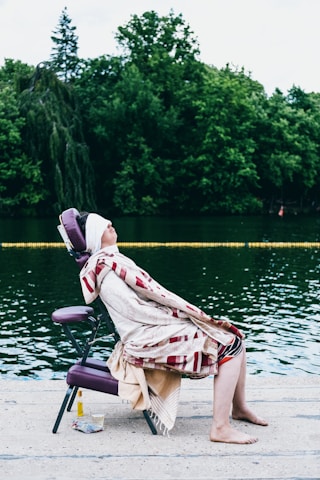 A person is seated on a purple portable massage chair at the edge of a body of water, covered with a patterned blanket, eyes closed and face slightly elevated. The background features lush green trees, and there is a small cloth and a bottle placed nearby on the ground.