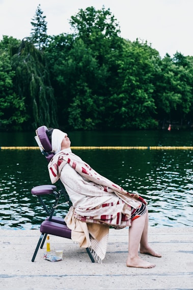 A person is seated on a purple portable massage chair at the edge of a body of water, covered with a patterned blanket, eyes closed and face slightly elevated. The background features lush green trees, and there is a small cloth and a bottle placed nearby on the ground.