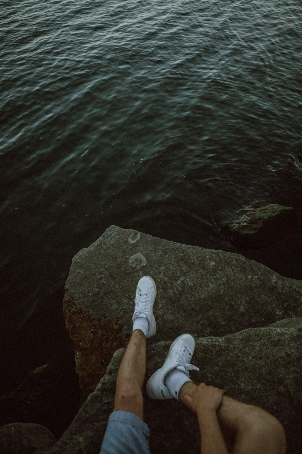 person in white shoes sitting on rock near body of water during daytime