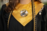 A close-up of a graduation gown with a gold stole that has an emblem at the center. Two colored cords, one blue and one gold, are draped over the gown. The gown also features a patch on the right side.
