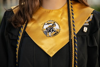 A close-up of a graduation gown with a gold stole that has an emblem at the center. Two colored cords, one blue and one gold, are draped over the gown. The gown also features a patch on the right side.