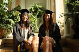 Two graduates sitting on steps, surrounded by lush green plants. They are wearing black graduation gowns and caps, smiling for the photo. The sunlight creates a warm and inviting atmosphere.