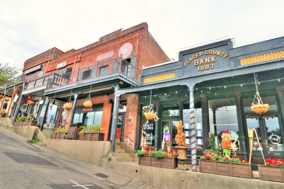 The image depicts a street view of several vintage-style buildings. The building on the right is labeled 'Placer County Bank 1881'. The facade is dark gray with yellow accents, and it features a large glass display window with mannequins and hanging baskets. To the left are more buildings with red brick exteriors and balconies. They are decorated with plants and also have large glass windows displaying various items. The street is sloped, and it appears to be a somewhat quaint and charming part of town.